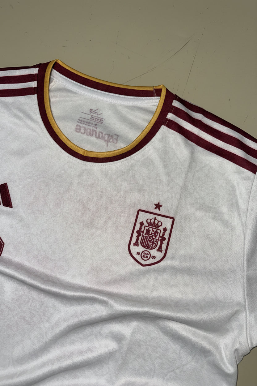 A close-up of a white Spain soccer jersey with maroon and gold trim, featuring the Spain crest on the chest and Adidas three-stripes on the shoulder.