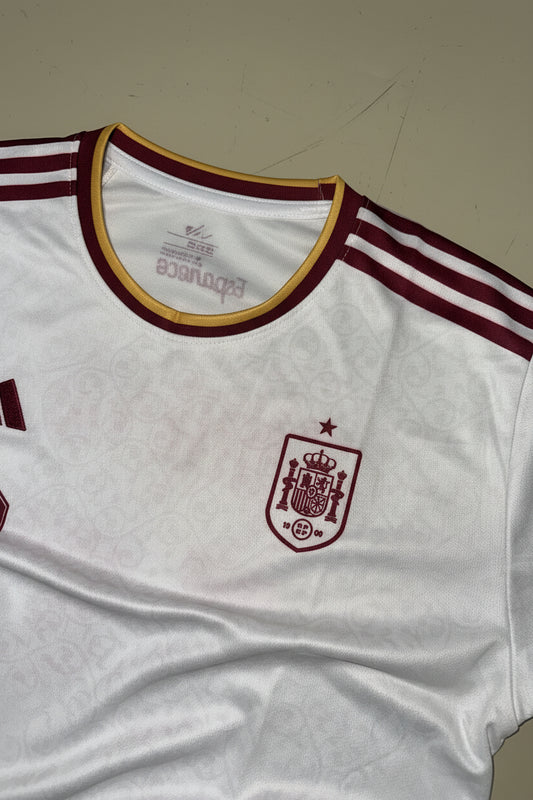 A close-up of a white Spain soccer jersey with maroon and gold trim, featuring the Spain crest on the chest and Adidas three-stripes on the shoulder.